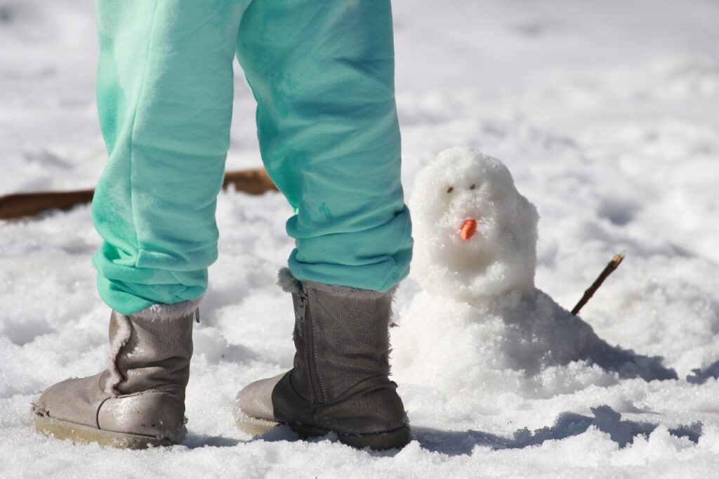 雪の上にブーツを履いて立つ。足元には小さな雪だるま。